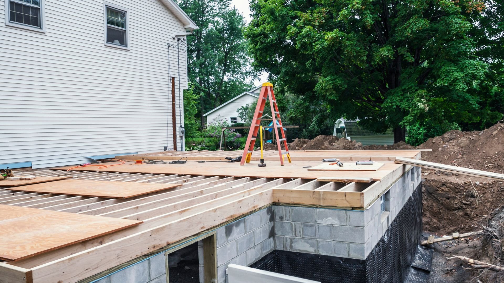 Wooden deck construction with orange ladder on concrete foundation near house