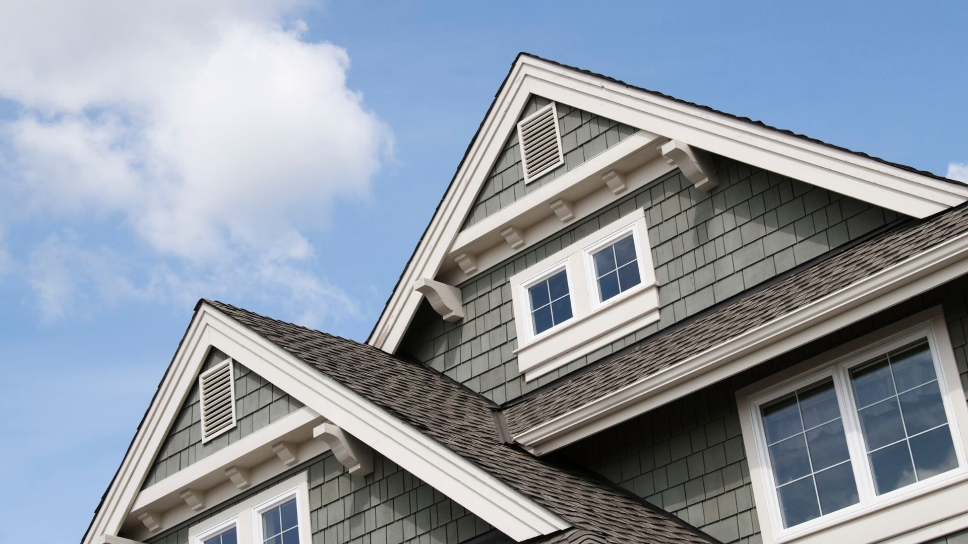 Gray shingled house with white trim and multiple windows against blue sky
